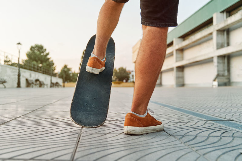 Unrecognizable talented male in sneakers riding shabby skateboard along street at sunset in summer