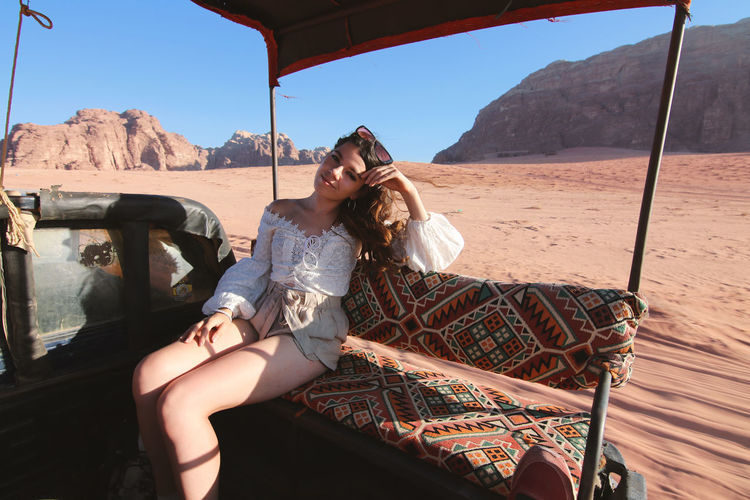 Woman sitting on car riding through desert against mountains