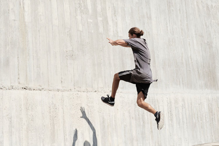 Side view of courageous young male jumping on concrete wall of building while performing dangerous stunt and doing parkour in city