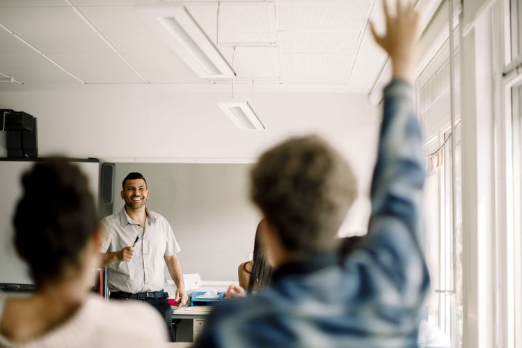 Male student with hand raised while smiling tutor standing in classroom