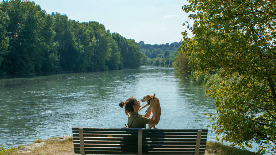 Side view of young woman carrying puppy while sitting on bench by lake against trees