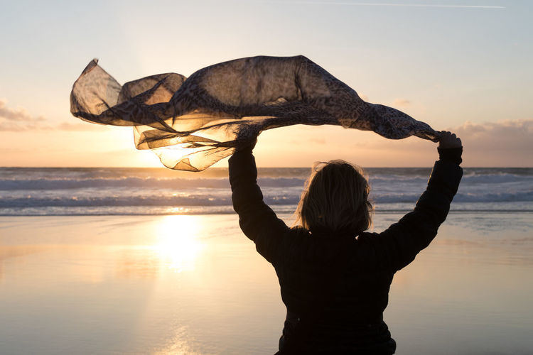 Rear view of woman holding scarf while standing at beach against sky