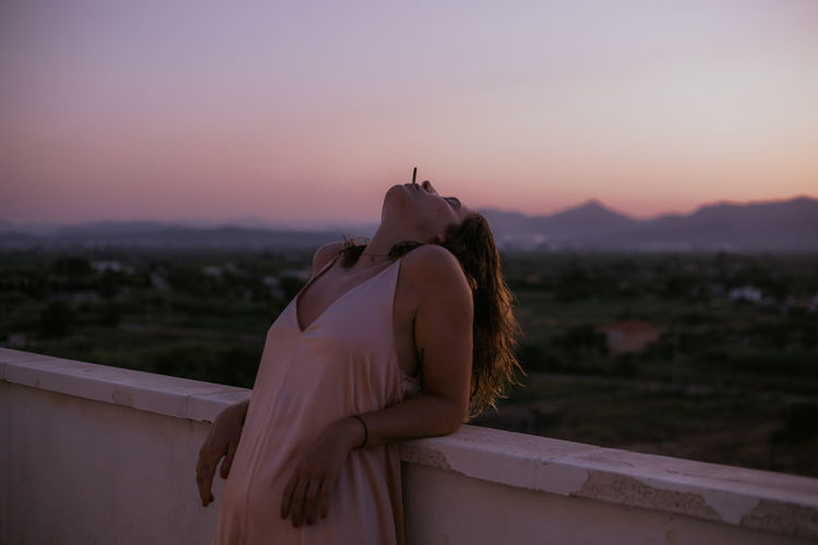 Side view of peaceful lady lying with closed eyes on fence of balcony and smoking cigarette with sunset on blurred background