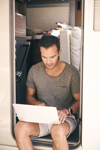 Mature man using laptop while sitting on doorway of travel trailer