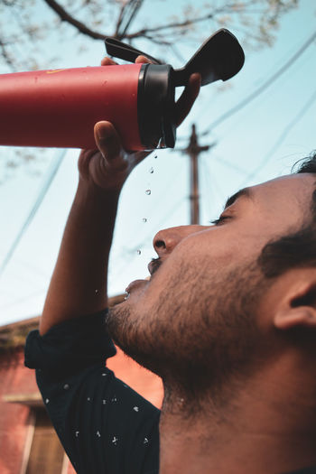 Close-up of young man pouring water on face against sky