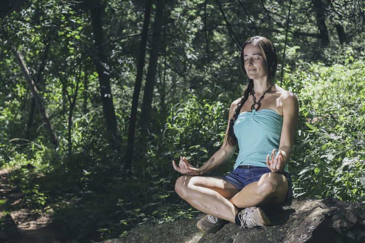 Beautiful woman sitting on a rock in meditation by tree in forest