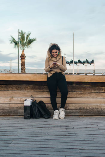 Satisfied curly haired young woman in casual wear smiling while using mobile phone on bench with shopping bags on modern quay
