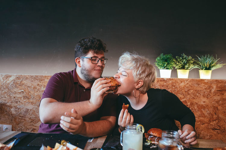 Smiling man feeding funny female friend with delicious burger while sitting at table in fast food cafe