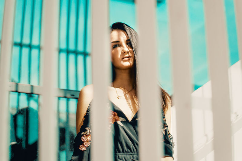 Portrait of smiling young woman seen through fence standing against building in city