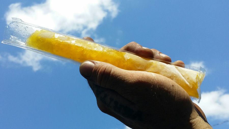 Cropped image of hand holding mango flavored popsicle against sky