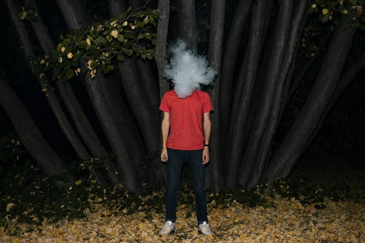Young man with smoke covering face standing against tree trunk in forest
