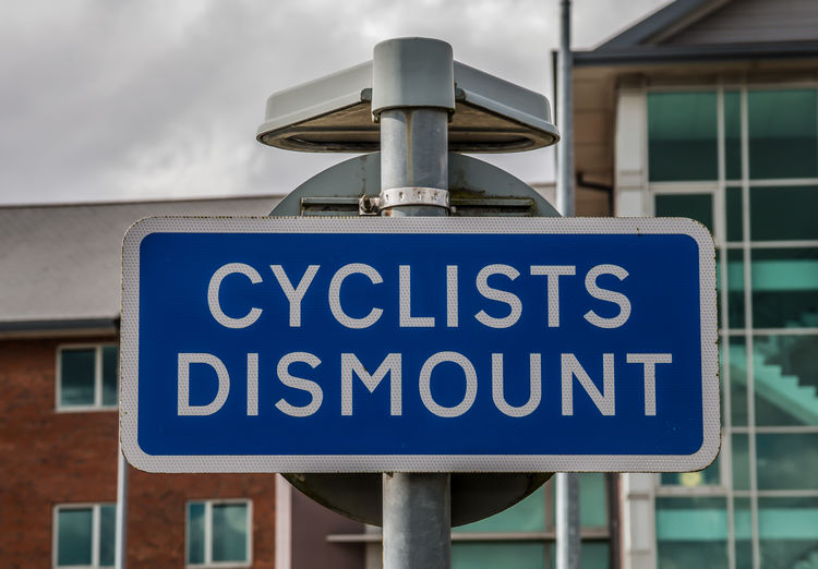 A bright blue sign with white writing advising cyclists dismount with office behind
