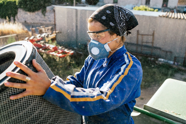 Side view of female farmer in protective goggles and respirator mask checking and cleaning filter of agricultural vehicle while working in farm yard