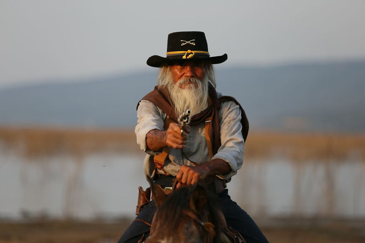 Portrait of man riding horse while holding handgun against sky