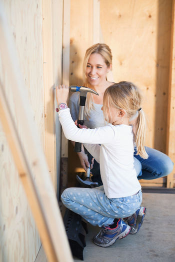 Happy mother looking at daughter hammering nail outside house being renovated