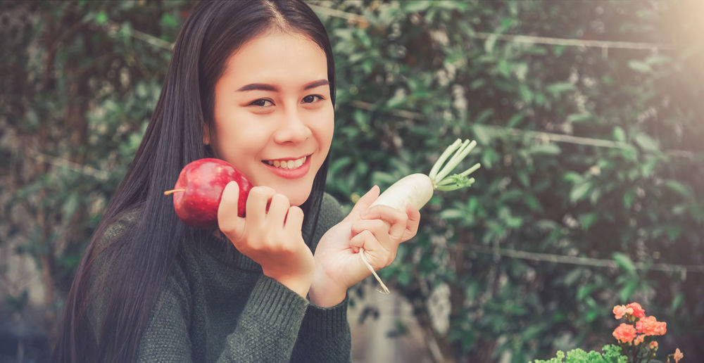 Portrait of smiling woman holding apple and radish against plants