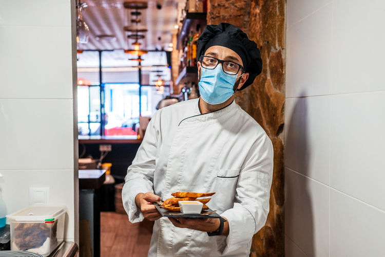 Serious male chef in uniform and medical mask standing with delicious snacks on slate board in cafe and looking at camera