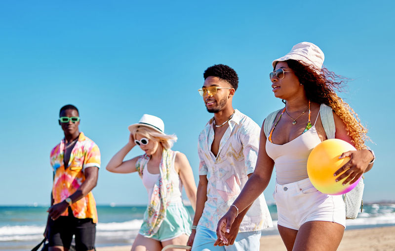 Full body smiling multiracial men and women in sunglasses and hat strolling on sand against cloudless blue sky while spending time on beach on summer day