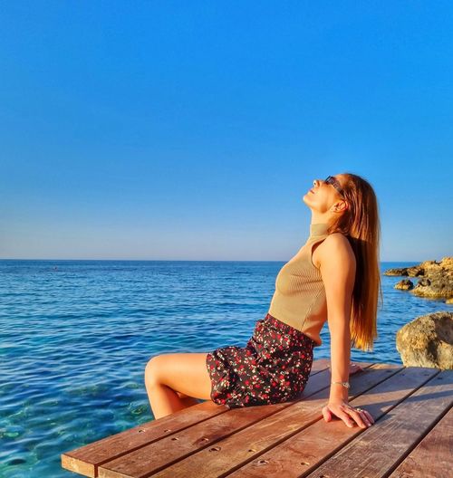 Young woman sitting on pier at beach against clear blue sky