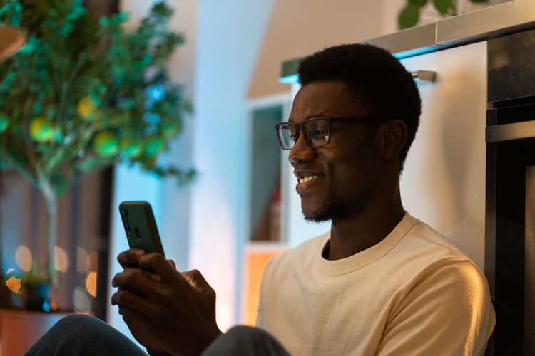 Young happy black guy using smartphone while relaxing and spending leisure time at cozy home