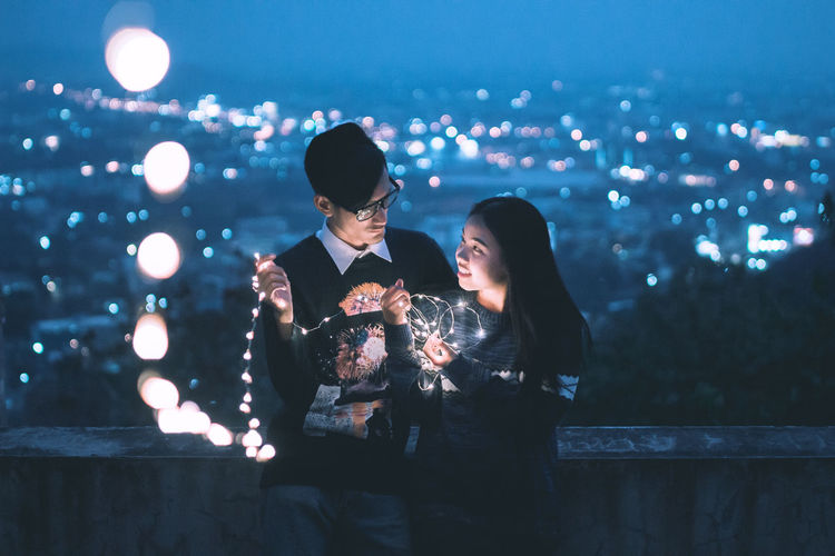 Couple holding illuminated string lights while standing in terrace at night
