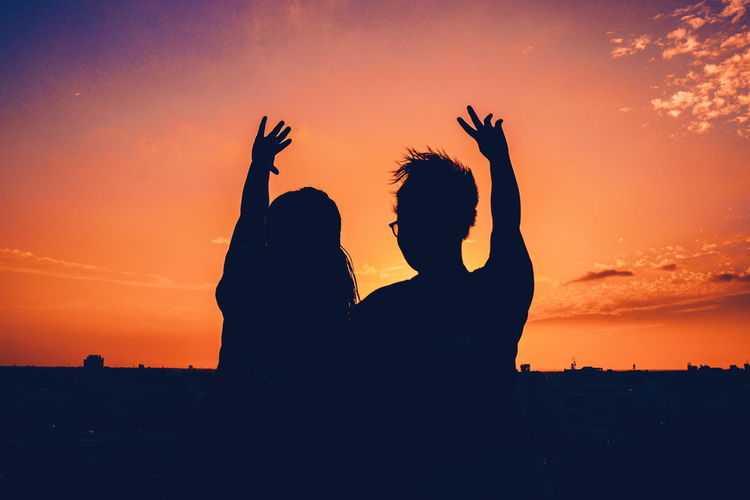 Silhouette friends with arms raised against sky during sunset