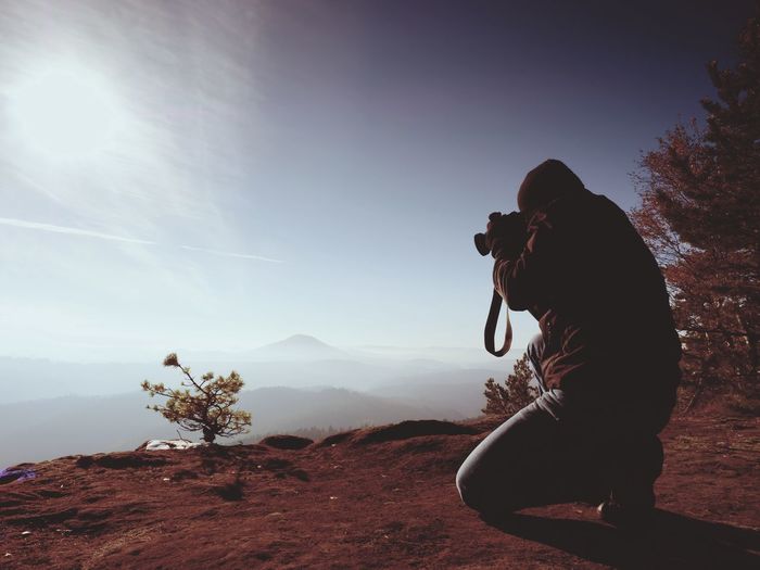 Photographer down on knees takes photos with mirror camera on peak of rock. dreamy landscape