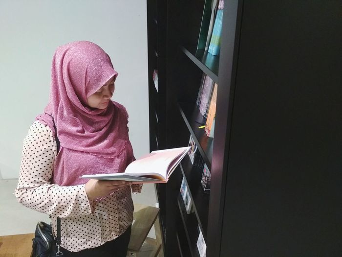 Woman reading book by bookshelf at library