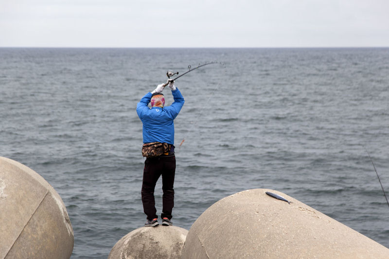 Rear view full length of person fishing while standing on rock against sea