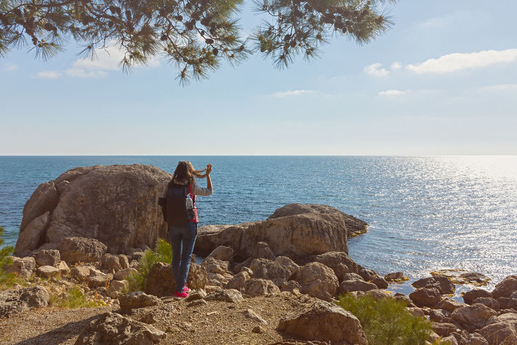 Rear view of woman sitting on rock by sea against sky