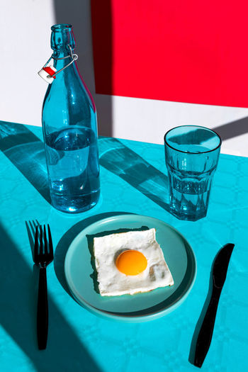 From above of composition of served fresh fried egg on plate with knife and fork opposite bottle of water and glass placed on blue table against red background in sunlight