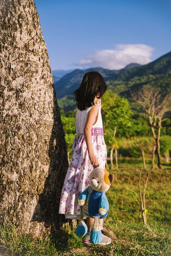 Girl holding toy while standing on grass by tree trunk against mountain and sky
