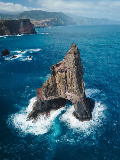 A rock formation standing out in the ocean next to the cliffs of madeira.