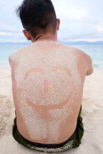 Rear view of shirtless man with anthropomorphic smiley face on his back at beach against sky
