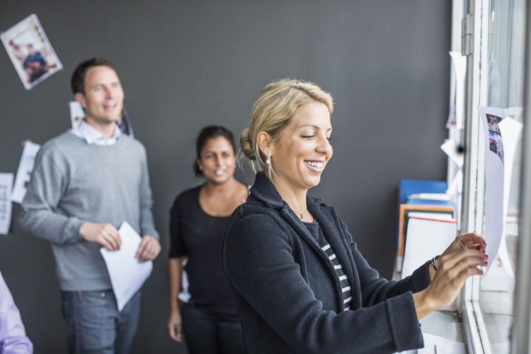 Mid adult businesswoman sticking paper with colleagues in background at office