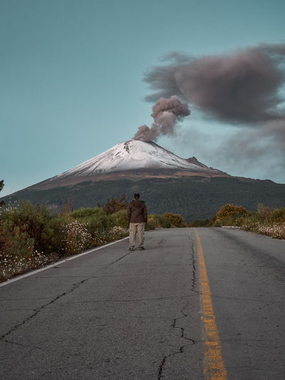 Rear view of man on road against smoke emitting from volcano and sky