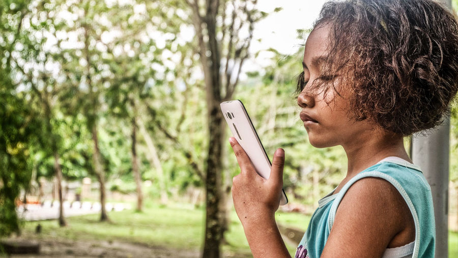 Side view of girl holding mobile phone against trees