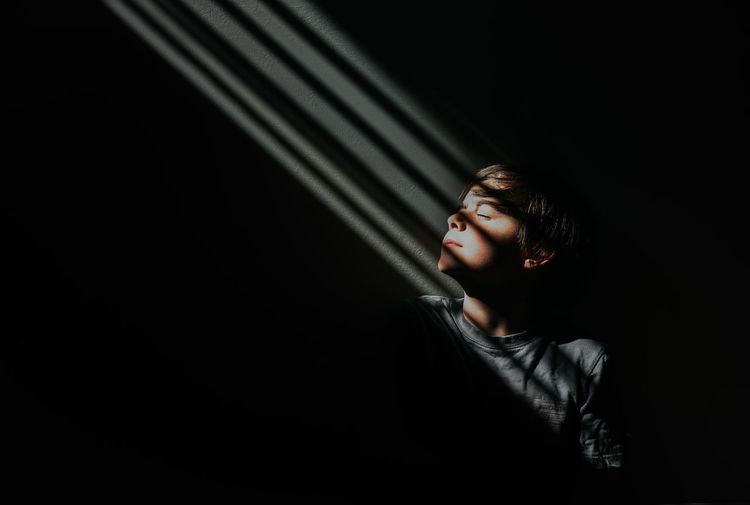 Young boy sitting in a patch of patterned light in a dark room.