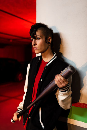 Contemporary female in bomber jacket looking away while leaning on wall with black baseball bat on shoulder with red light on background