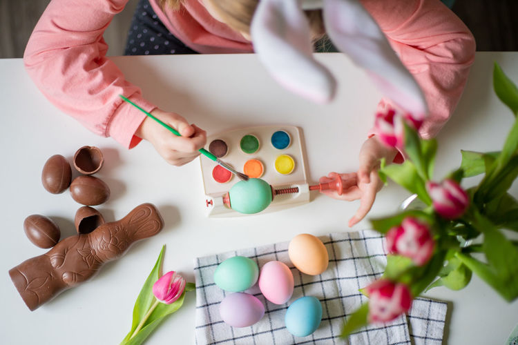 Top view of a little girl with bunny ears holding paintbrush and decorating easter eggs watercolors.