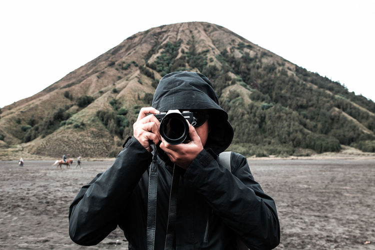 Young man photographing with digital camera against mountain
