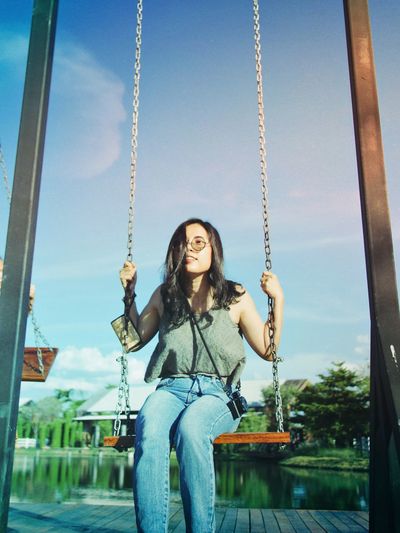 Woman sitting on swing in playground against lake and sky
