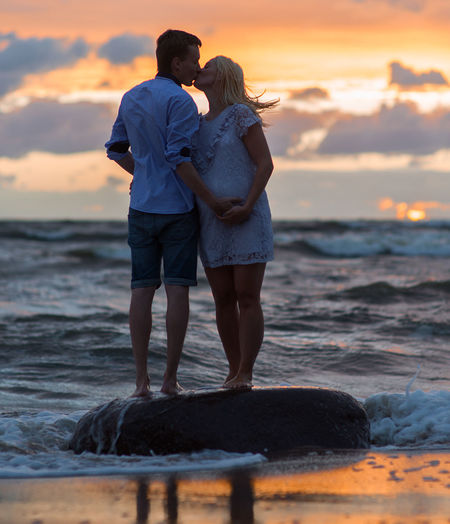 Expectant couple kissing while standing on shore at beach during sunset