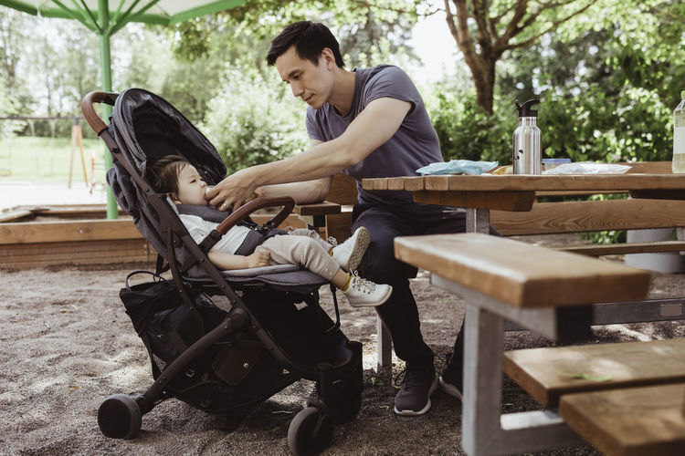 Father feeding baby son while sitting in playground