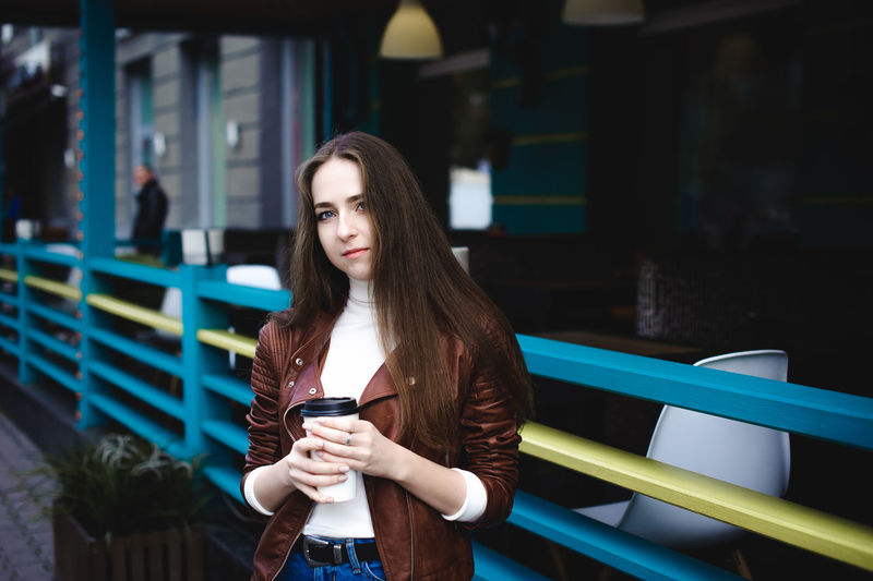 Portrait of young woman holding disposable cup while standing by railing