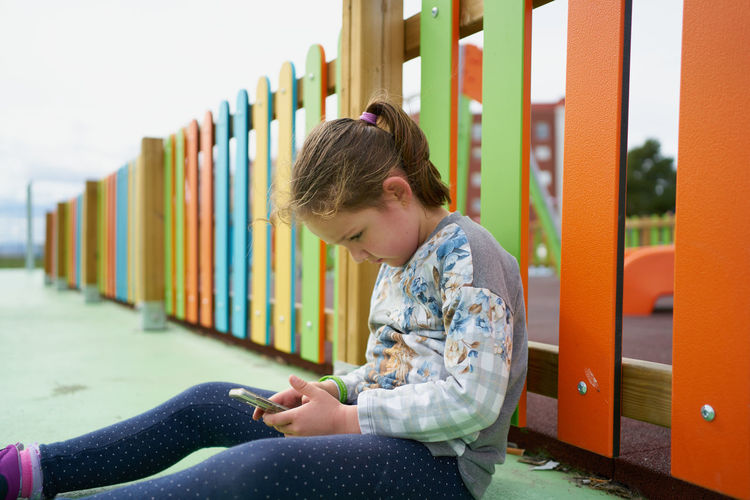 Side view of little girl sitting on playground and browsing mobile phone