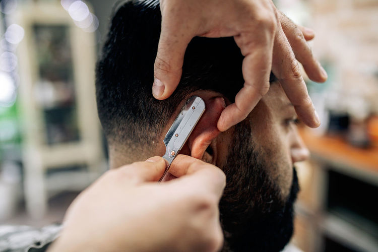Crop anonymous barber using sharp straight blade for cutting hair of male customer in professional barbershop