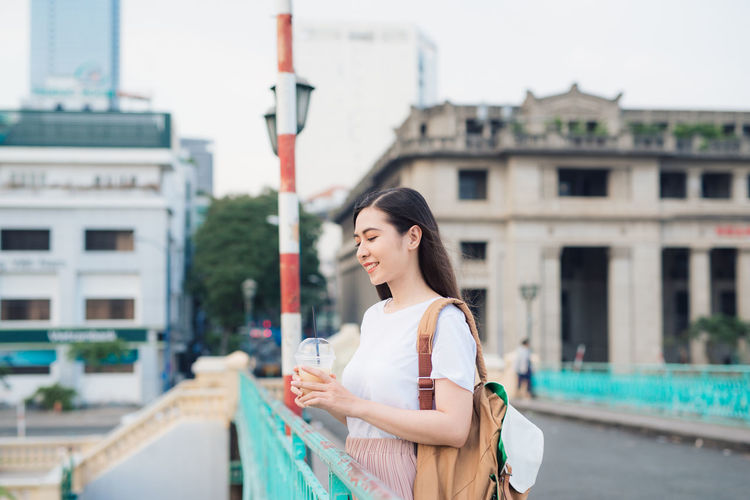 Smiling beautiful woman holding drink while standing on bridge