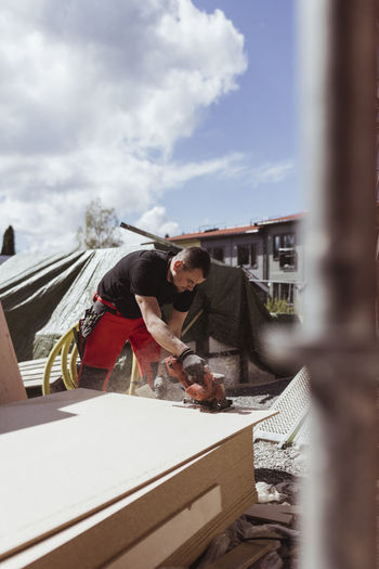 Male carpenter cutting wooden plank from electric saw at construction site during sunny day