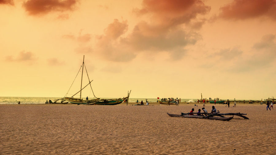 People sitting on sailboats at beach against sky during sunset